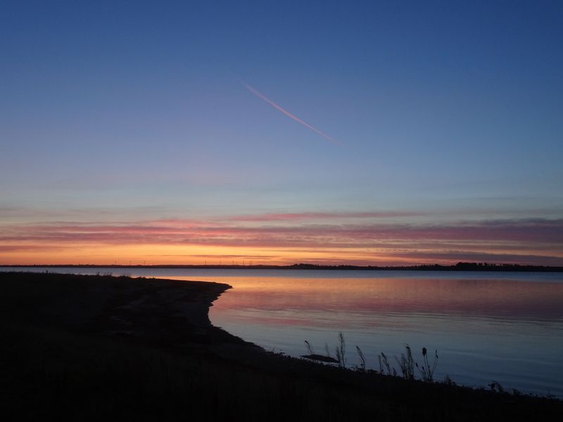 Roze en oranje zonsopkomst boven stil water, met een condensstreep in de lucht